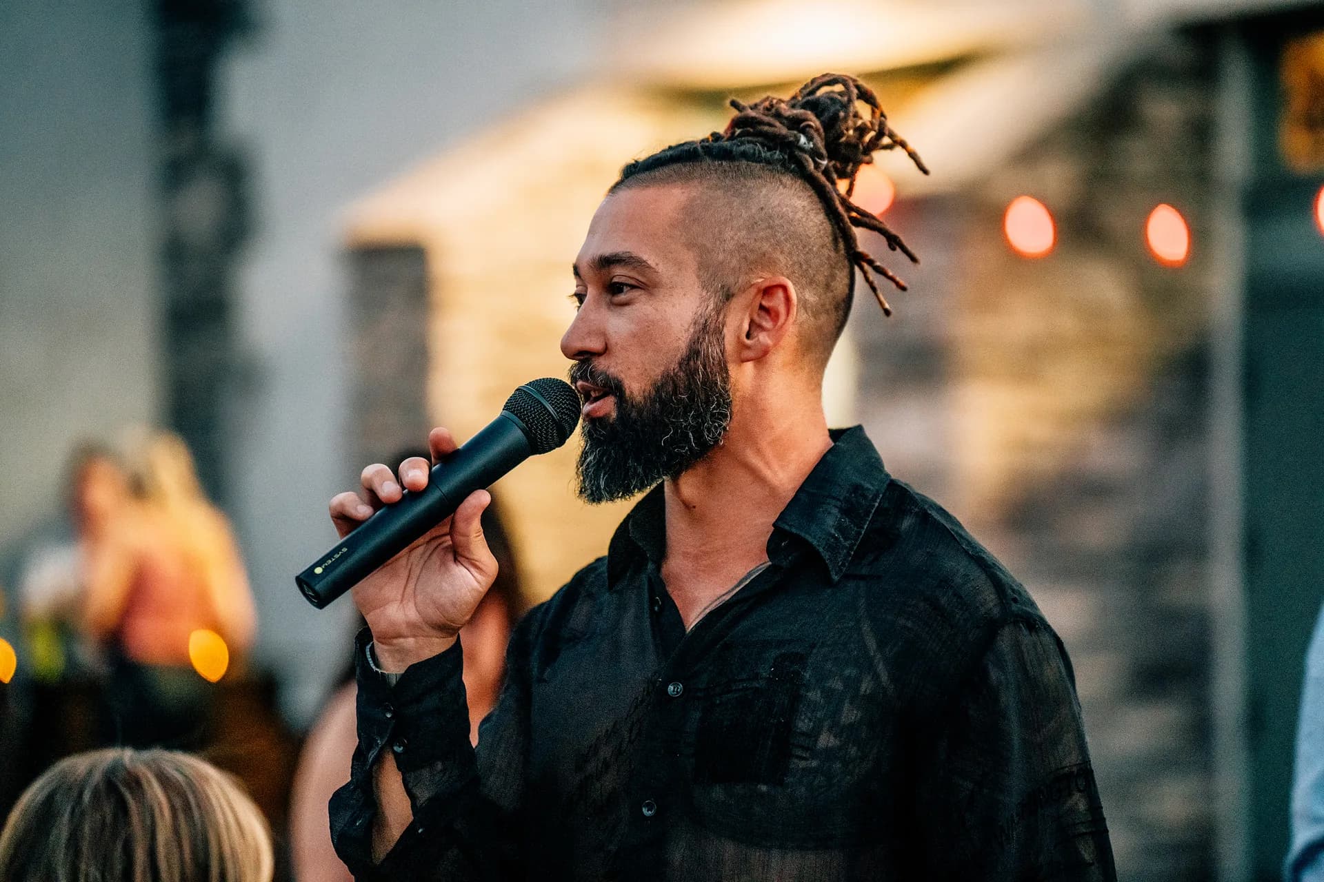 A bearded man with a shaved-sided top-knot holds a handheld microphone close to his mouth, string lights glowing behind him in the evening.