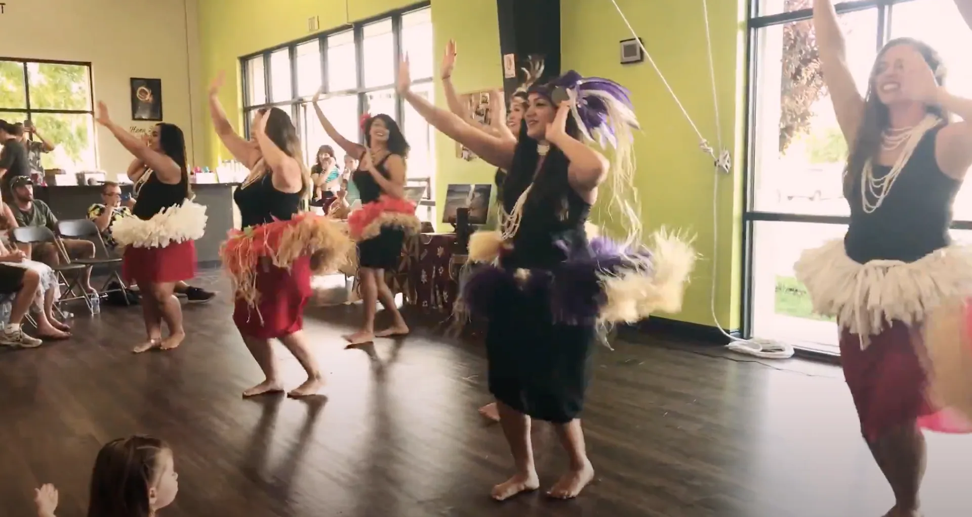A row of women in traditional grass skirts and leis dance with raised arms in a bright studio.
