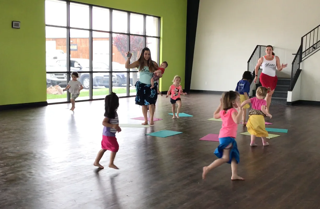 Small children and their mothers dance barefoot on colorful mats inside a sunlit dance studio.