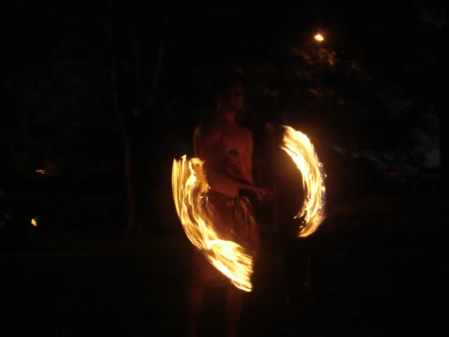 A shirtless figure spins two fire poi in glowing arcs beneath dark trees at night.