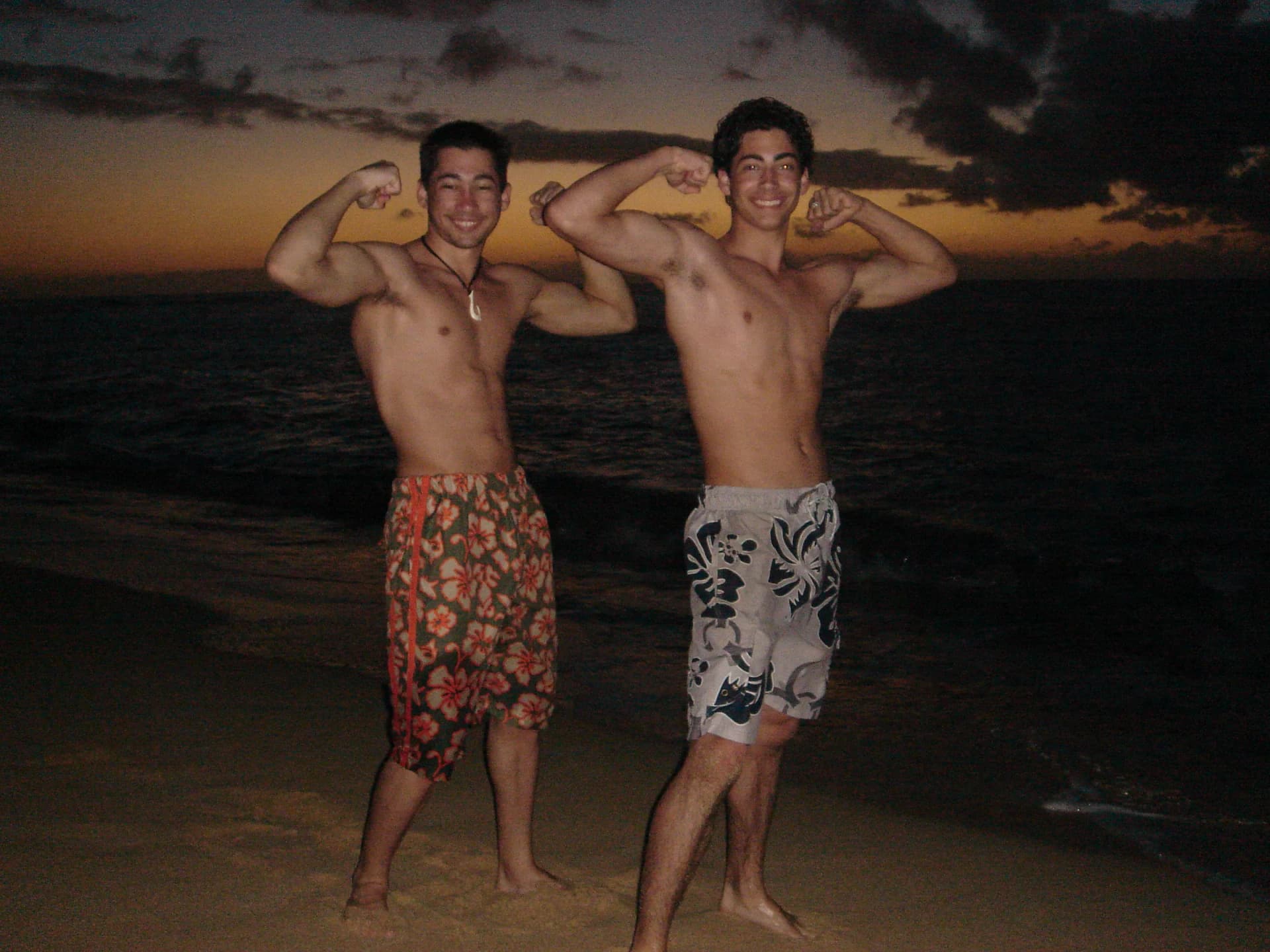 Two young men flex their muscles and smile on a beach at dusk, ocean behind them.