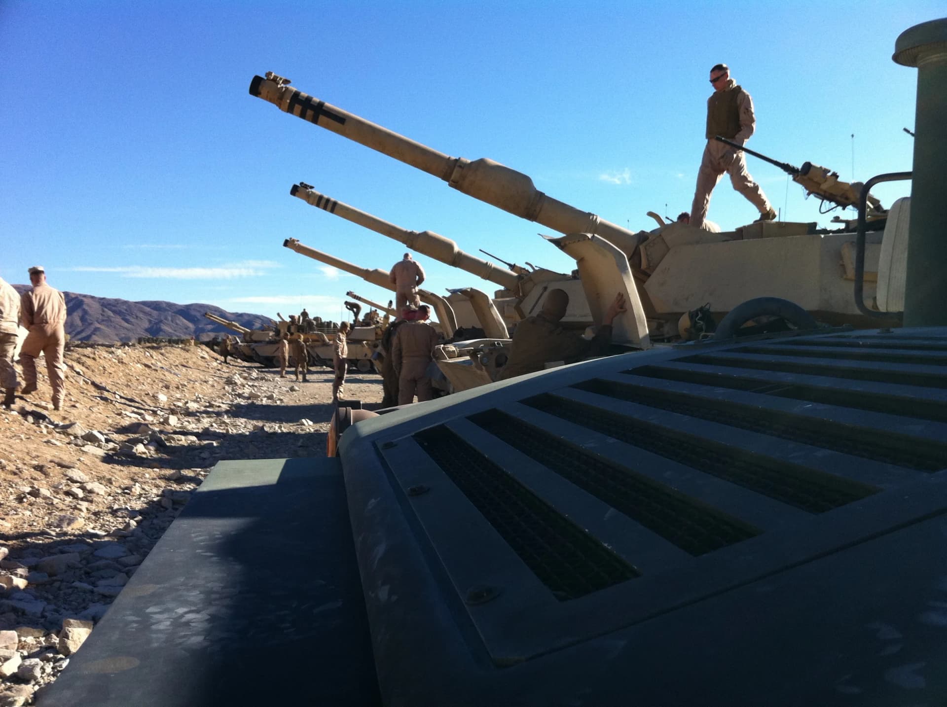 Marines in desert camouflage work around a row of howitzer cannons under a clear blue sky.