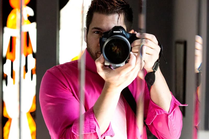 A man in a pink shirt holds a camera up to a mirror, capturing his reflection.