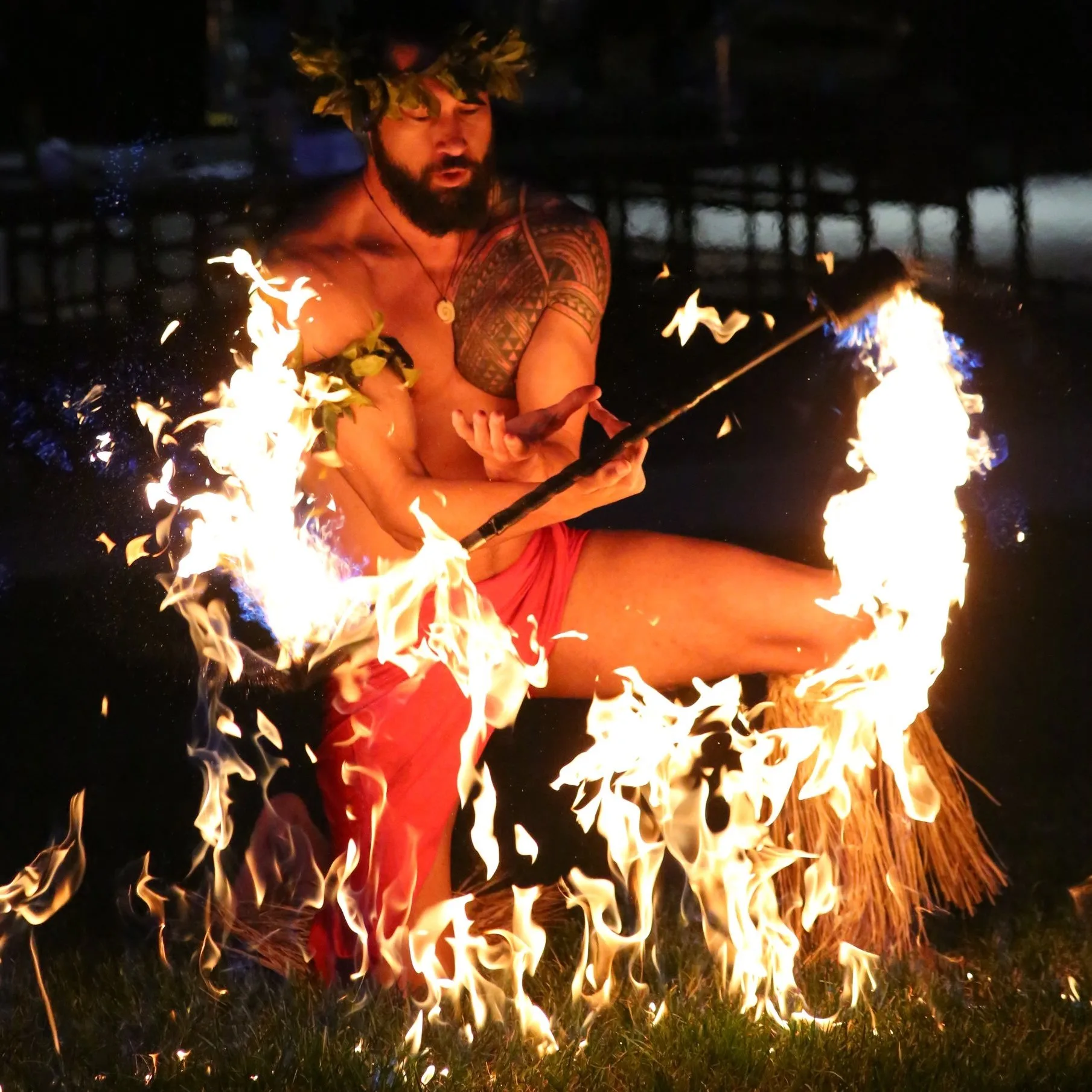A tattooed fire performer in a red wrap crouches low, flames circling his kicked-out leg.