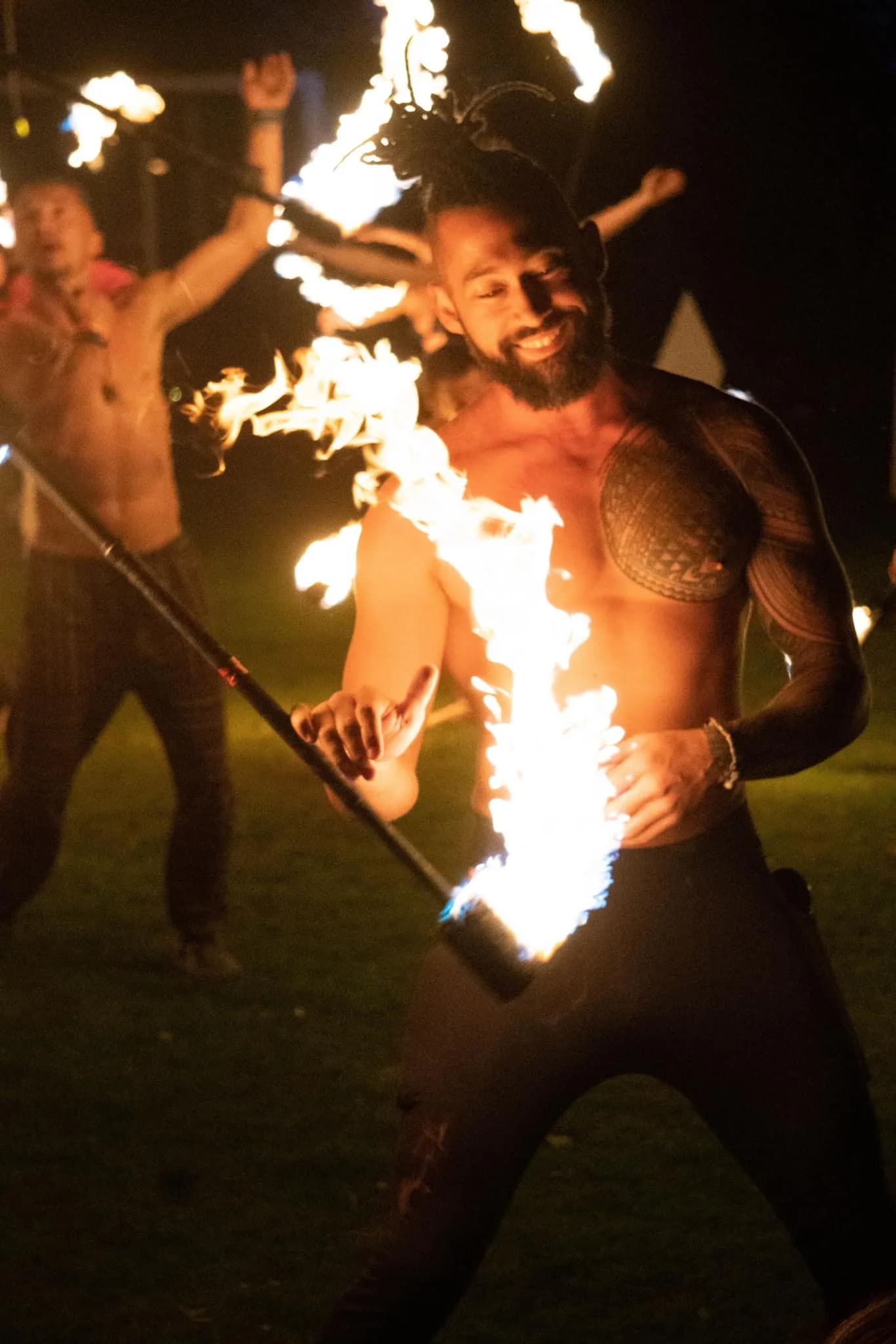 A smiling fire performer with a tattooed chest swings a flaming torch as others fire dance behind him.