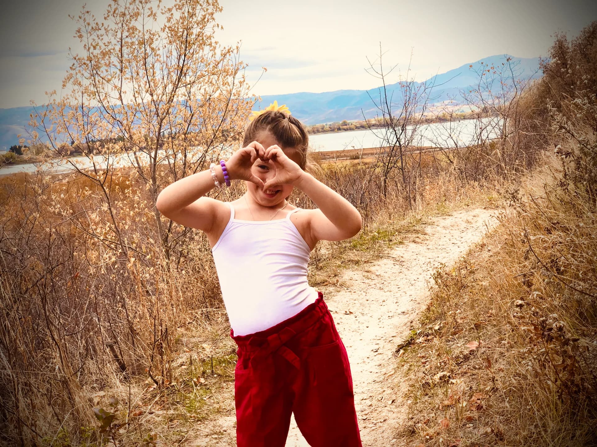 A young girl forms a heart shape with her hands on a trail overlooking a lake and mountains.